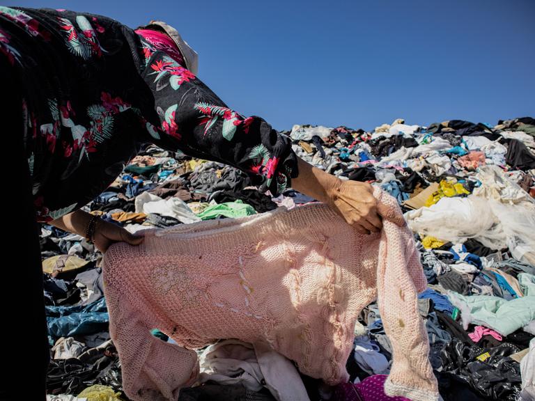 Eine Frau sucht unter einem Berg gebrauchter Kleidern in einer Müll-Deponie ein Kleidungsstück. In der nahe gelegenen Freihandelszone von Iquique kamen 2021 bis Oktober 29.178 Tonnen gebrauchter Kleidung an.