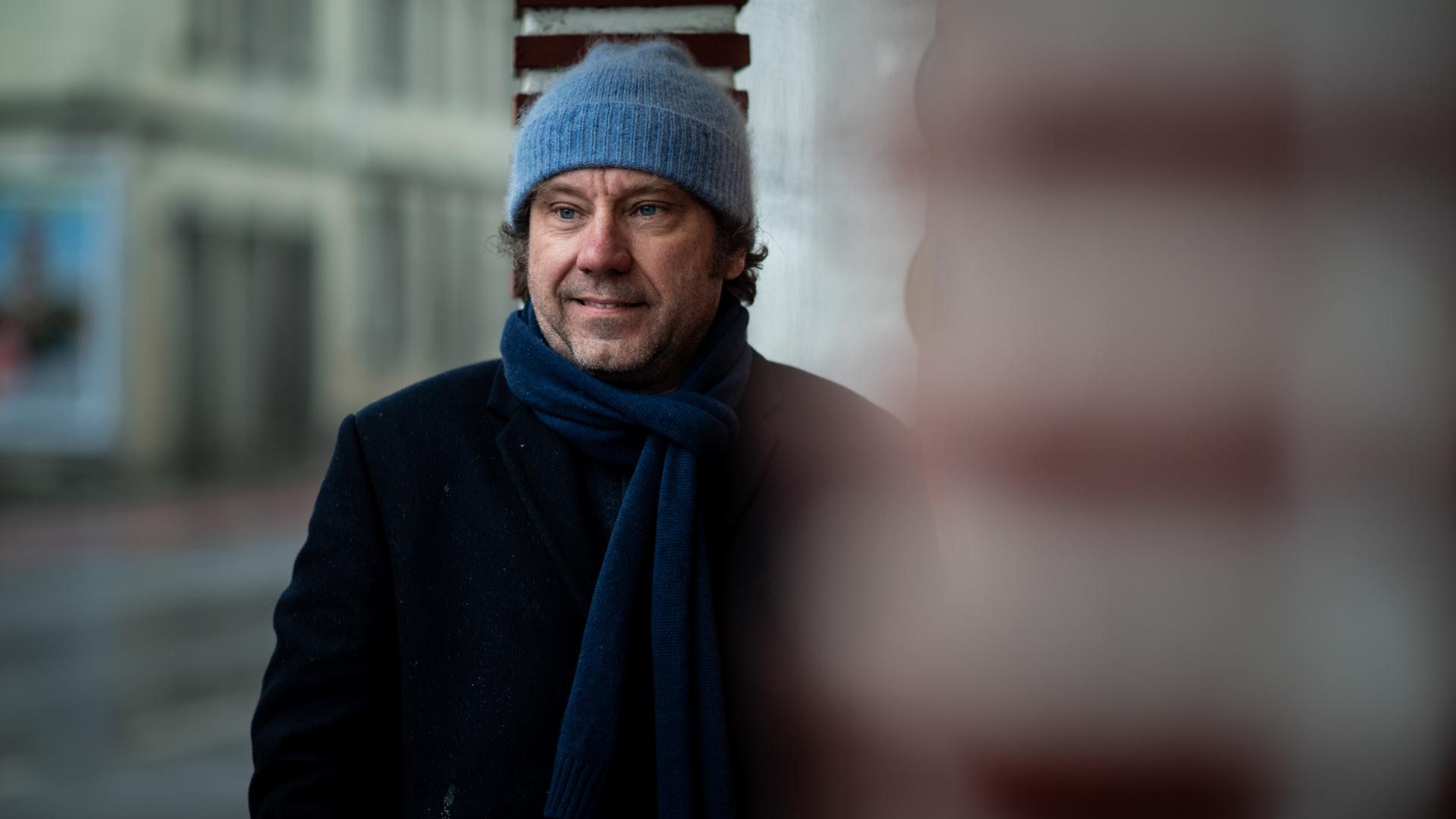 Gregor Sander steht vor einer Säule, er trägt eine hellblaue Wollmütze und einen dunkelblauen Schal und schaut lächelnd zur Seite. Gregor Sander steht vor einer Säule, er trägt eine hellblaue Wollmütze und einen dunkelblauen Schal und schaut lächelnd zur Seite.