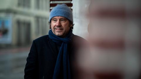 Gregor Sander steht vor einer Säule, er trägt eine hellblaue Wollmütze und einen dunkelblauen Schal und schaut lächelnd zur Seite. Gregor Sander steht vor einer Säule, er trägt eine hellblaue Wollmütze und einen dunkelblauen Schal und schaut lächelnd zur Seite.