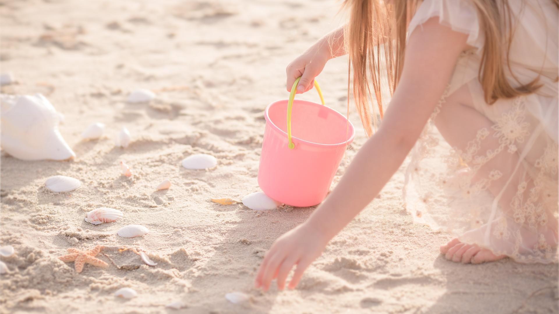 Ein Kind mit rosa farbener Kleidung und einem rosa Eimer mit gelbem Henkel hockt an einem Strand und sammelt Muscheln.