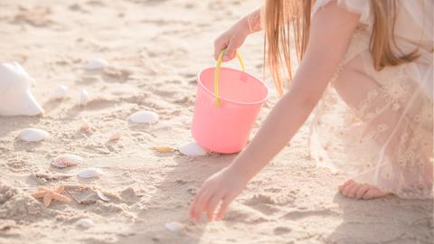 Ein Kind mit rosa farbener Kleidung und einem rosa Eimer mit gelbem Henkel hockt an einem Strand und sammelt Muscheln.