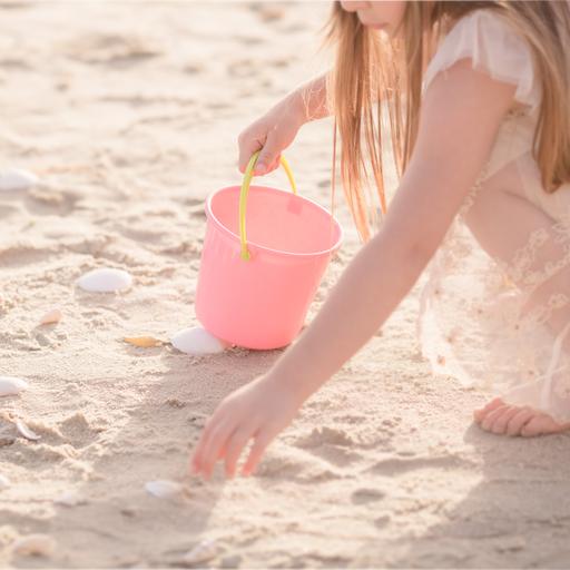 Ein Kind mit rosa farbener Kleidung und einem rosa Eimer mit gelbem Henkel hockt an einem Strand und sammelt Muscheln.