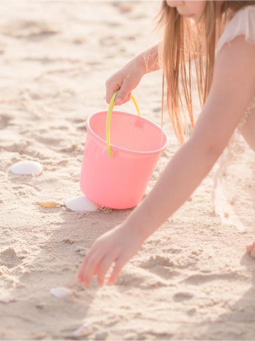 Ein Kind mit rosa farbener Kleidung und einem rosa Eimer mit gelbem Henkel hockt an einem Strand und sammelt Muscheln.