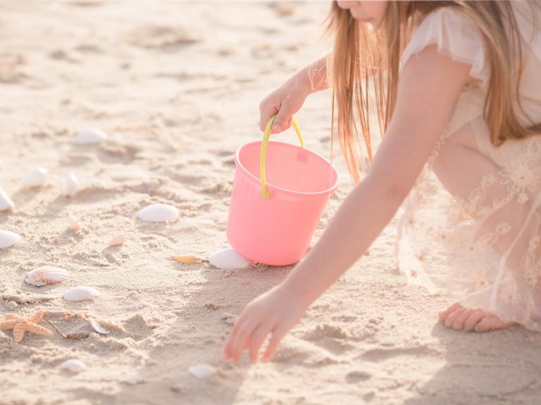 Ein Kind mit rosa farbener Kleidung und einem rosa Eimer mit gelbem Henkel hockt an einem Strand und sammelt Muscheln.