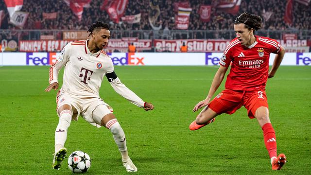 Ein Spieler von dem Verein Bayern München hat den Ball. Ein Spieler von dem Verein Benfica Lissabon steht neben ihm und holt mit dem Bein aus.