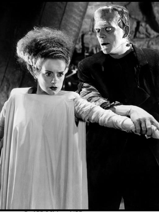 Das Schwarzweißfoto zeigt eine Frau in heller Kleidung mit starrem Blick und eine männliche Person mit weißem Auge und verformten Kopf, der hinter ihr steht