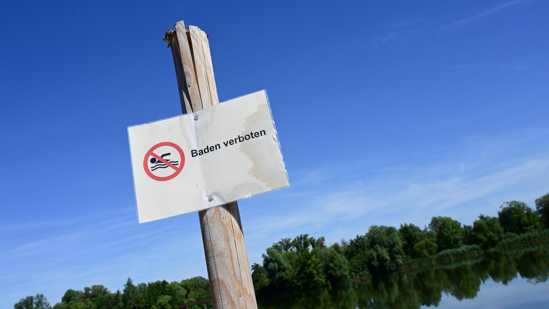 An einem Holzpfahl ist ein Schild befestigt. Darauf steht "Baden verboten". Im Hintergrund sind ein See und das Ufer zu erkennen. An einem Holzpfahl ist ein Schild befestigt. Darauf steht "Baden verboten". Im Hintergrund sind ein See und das Ufer zu erkennen.