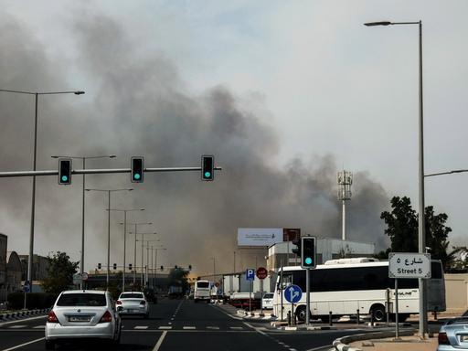 Autos fahren in Doha auf einer Straße, im Hintergrund sieht man Rauch aufsteigen.