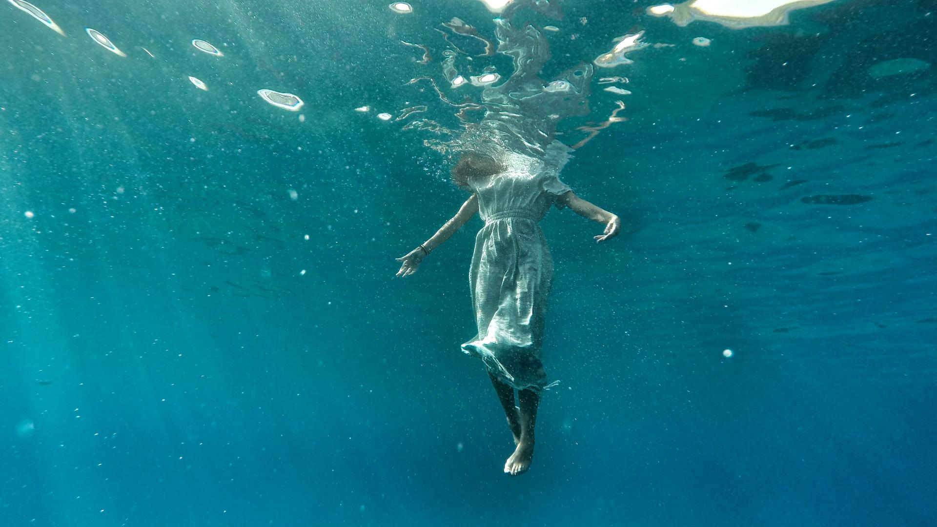 Blick von unter Wasser: Wir sehen den Körper einer Frau aufrecht im Wasser schweben, nur der Kopf ist oberhalb der Wasseroberfläche und daher beim Betrachten des Fotos nicht sichtbar.