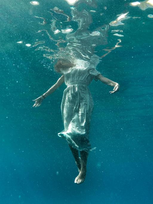 Blick von unter Wasser: Wir sehen den Körper einer Frau aufrecht im Wasser schweben, nur der Kopf ist oberhalb der Wasseroberfläche und daher beim Betrachten des Fotos nicht sichtbar.