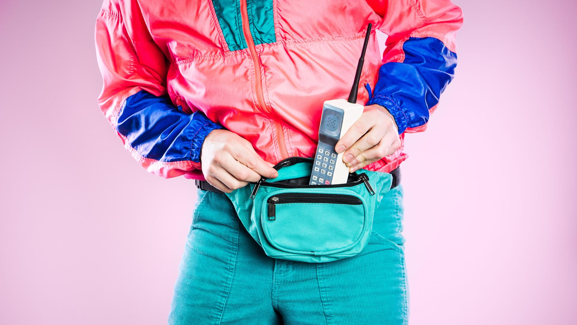Ein Mann, der bunte Kleidung aus den 1980er bis 1990er Jahren trägt, steckt ein altes Mobiltelefon in seine Gürteltasche, das den neuesten Stand von Stil und Technologie für diese Zeit repräsentiert.  