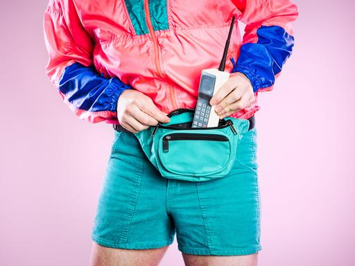 Ein Mann, der bunte Kleidung aus den 1980er bis 1990er Jahren trägt, steckt ein altes Mobiltelefon in seine Gürteltasche, das den neuesten Stand von Stil und Technologie für diese Zeit repräsentiert. Ein Mann, der bunte Kleidung aus den 1980er bis 1990er Jahren trägt, steckt ein altes Mobiltelefon in seine Gürteltasche, das den neuesten Stand von Stil und Technologie für diese Zeit repräsentiert.