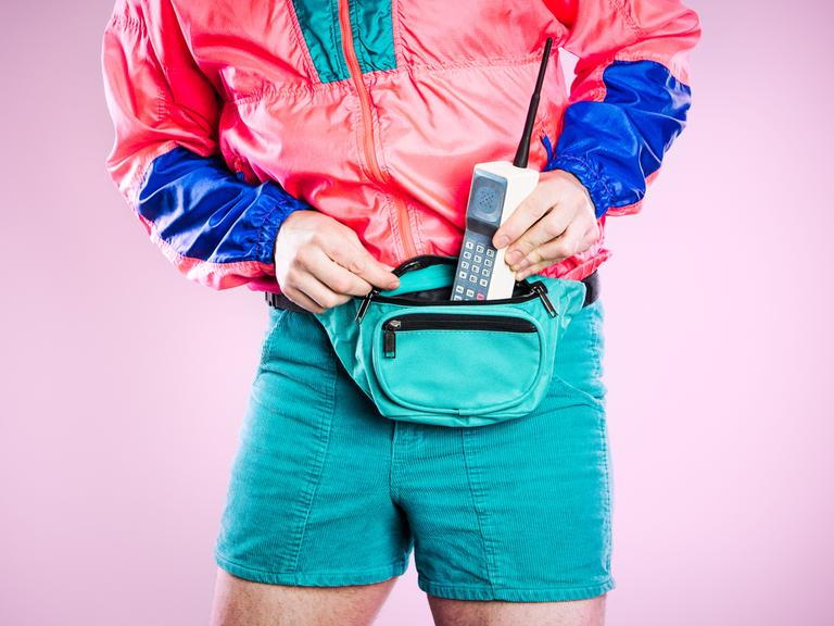 Ein Mann, der bunte Kleidung aus den 1980er bis 1990er Jahren trägt, steckt ein altes Mobiltelefon in seine Gürteltasche, das den neuesten Stand von Stil und Technologie für diese Zeit repräsentiert.  