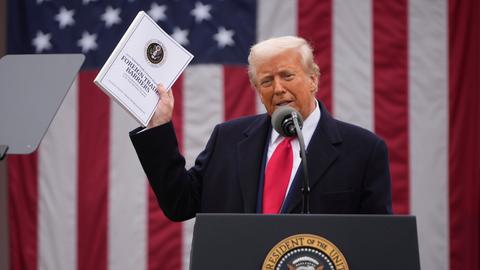 Donald Trump steht vor einer US-Flagge, spricht an einem Pult in ein Mikro und hält dabei eine dicke Broschüre hoch, auf der "Foreign Trade Barriers" steht.