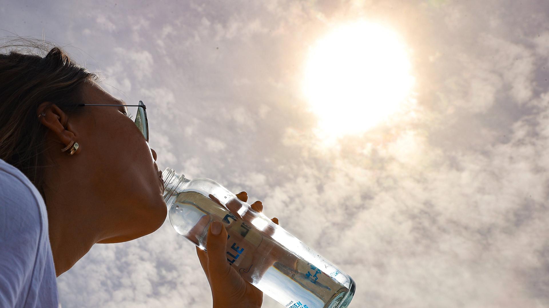 Eine Frau trinkt aus einer Flasche Wasser. Über ihr scheint die Sonne.
