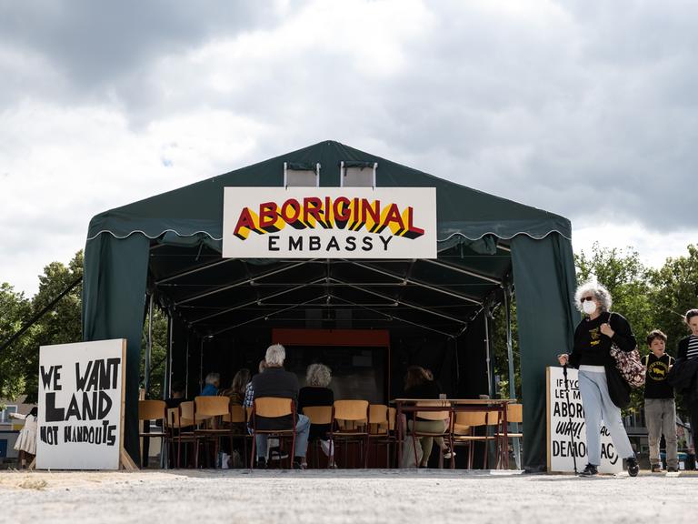 An einem großen Zelt hängt ein Schild  "Aboriginal Embassy", innen sitzen Besucher. 