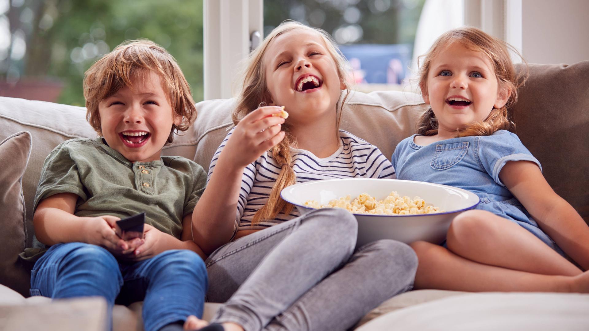 Drei Kinder sitzen lachend auf dem Sofa und schauen fern. Ein Mädchen hält eine große Schüssel mit Popcorn, ein Junge hält die Fernbedienung.