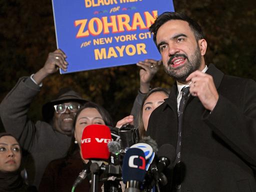 Zohran Mamdani spricht am 3. November 2025 in Astoria, Queens vor Anhängern in Mikrofone. Hinter ihm wird ein Schild mit der Aufschrift "Zohran for New York City Mayor"