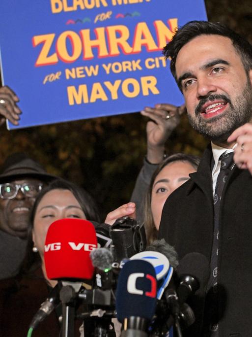 Zohran Mamdani spricht am 3. November 2025 in Astoria, Queens vor Anhängern in Mikrofone. Hinter ihm wird ein Schild mit der Aufschrift "Zohran for New York City Mayor"