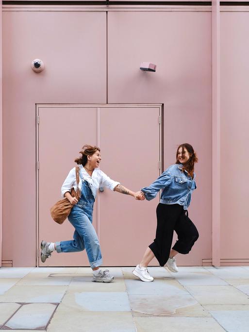 Zwei Frauen laufen Hand in Hand an einem hellrosa Gebäude vorbei.