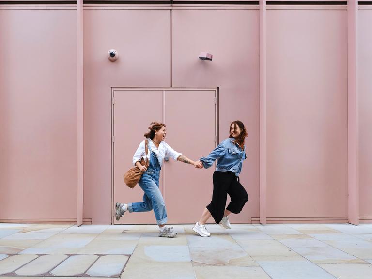 Zwei Frauen laufen Hand in Hand an einem hellrosa Gebäude vorbei.