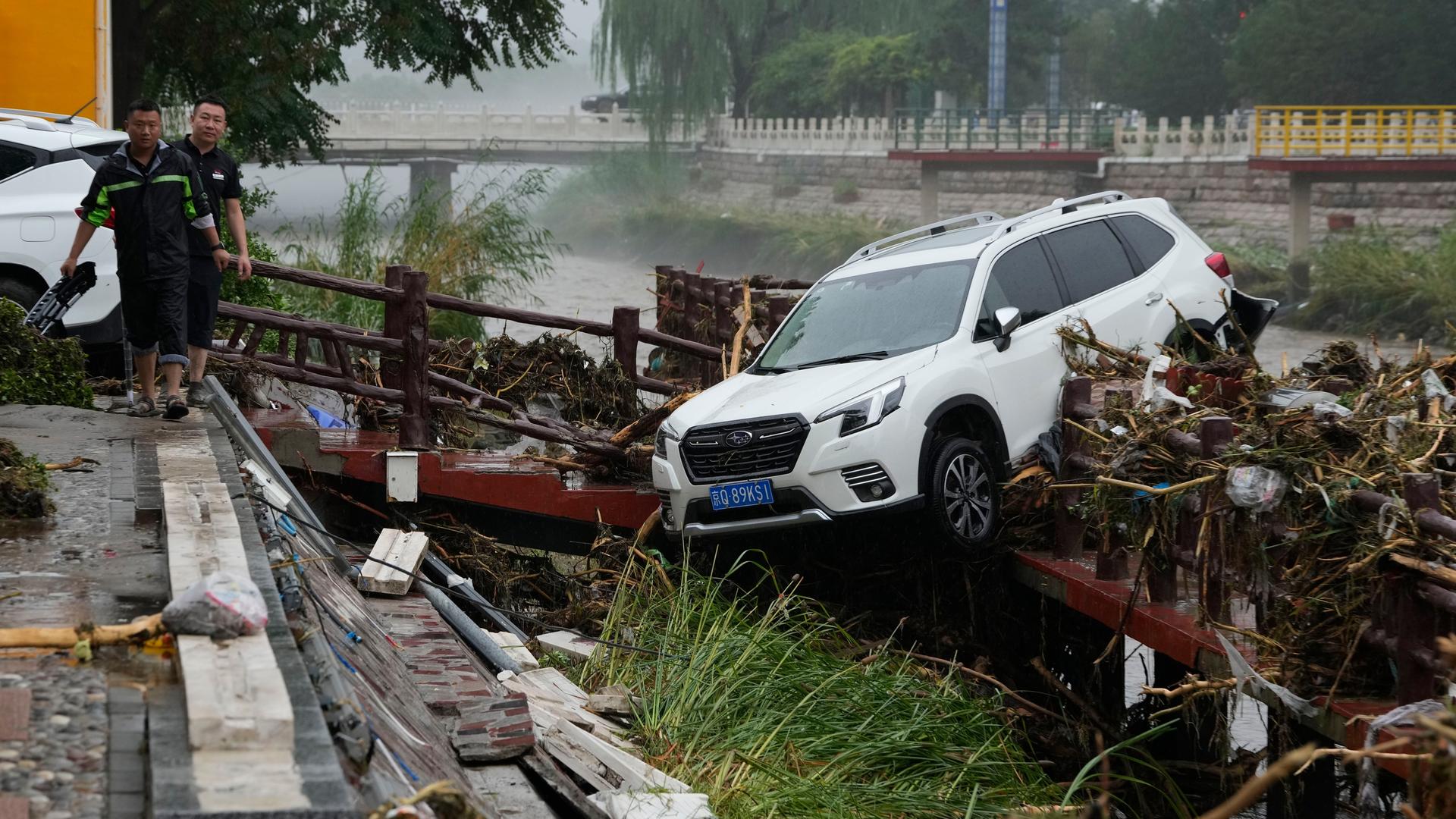 Anwohner gehen im chinesischen Bezirk Mentougou neben einem von den Fluten weggespülten Fahrzeug entlang.