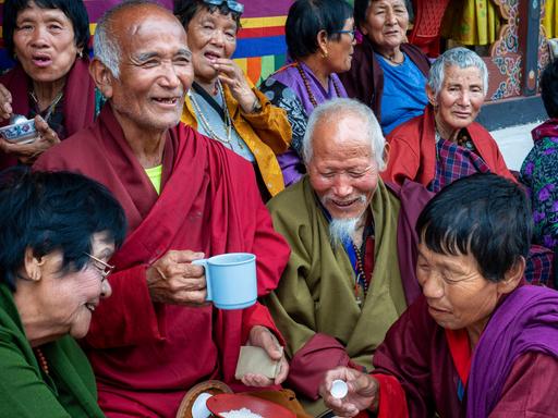 Gläubige Buddhisten in traditioneller Kleidung. Wollene Mäntel in Dunkelrot und Olivgrün. Die älteren Menschen unterhalten sich, viele lächeln. Ein Mann hält eine Tasse in der Hand. Alle machen einen zufriedenen Eindruck

