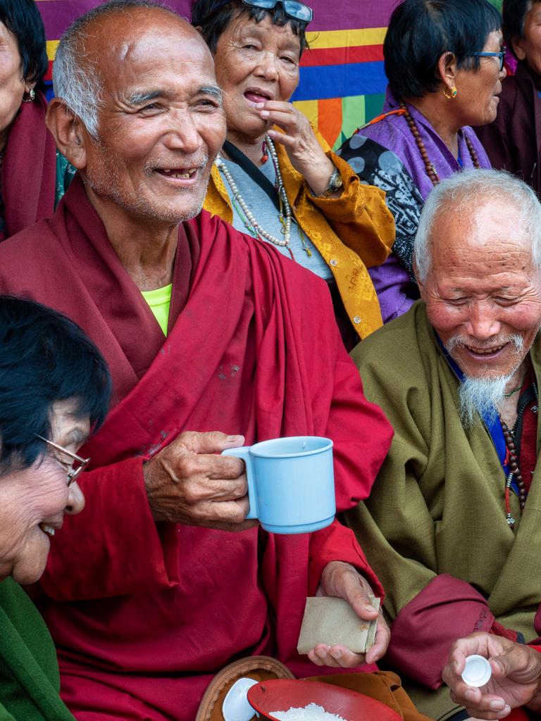 Gläubige Buddhisten in traditioneller Kleidung. Wollene Mäntel in Dunkelrot und Olivgrün. Die älteren Menschen unterhalten sich, viele lächeln. Ein Mann hält eine Tasse in der Hand. Alle machen einen zufriedenen Eindruck
