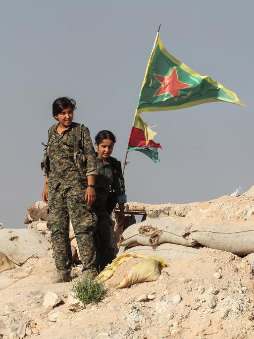 Zwei Frauen in Militäruniformen stehen auf einem Berg aus Schutt und Sandsäcken. Im Hintergrund weht die Flagge von Rojava und die der YPJ, eine Frauenverteidigungseinheit.
