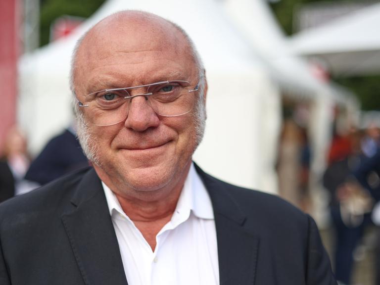 Ulrich Schneider mit weißen Haaren und Metallbrille trägt einen Anzug und schaut freundlich in die Kamera