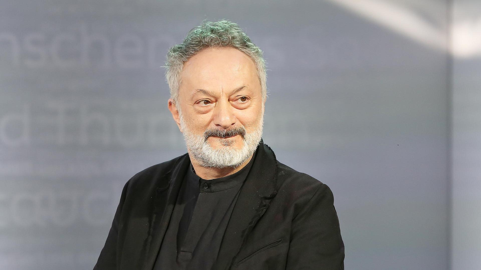 Feridun Zaimoglu sitzt vor einem grauen Hintergrund und guckt an der Kamera vorbei