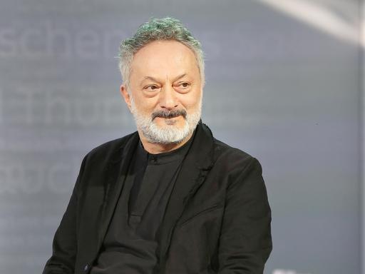 Feridun Zaimoglu sitzt vor einem grauen Hintergrund und guckt an der Kamera vorbei
