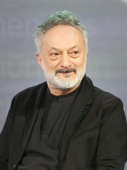 Feridun Zaimoglu sitzt vor einem grauen Hintergrund und guckt an der Kamera vorbei