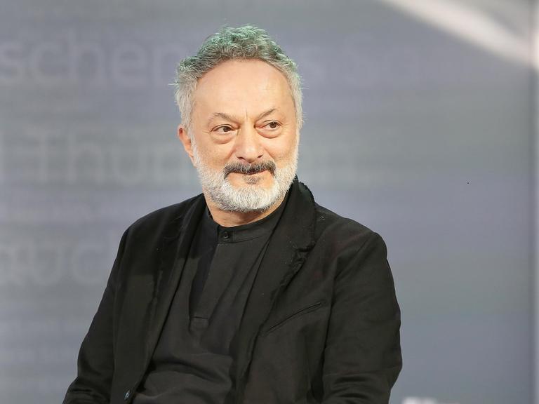 Feridun Zaimoglu sitzt vor einem grauen Hintergrund und guckt an der Kamera vorbei