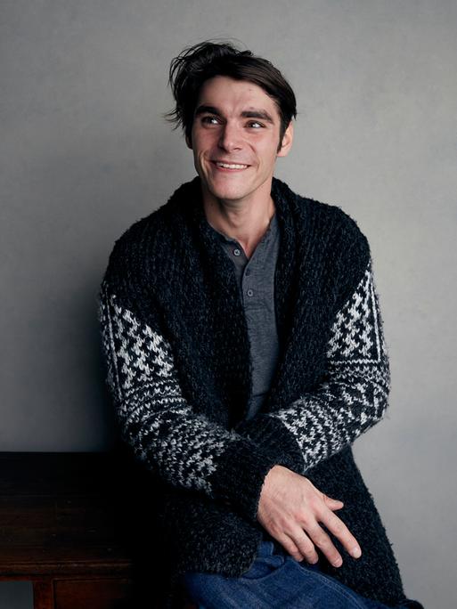 RJ Mitte sitzt auf einem Hocker vor einer grauen Wand. 