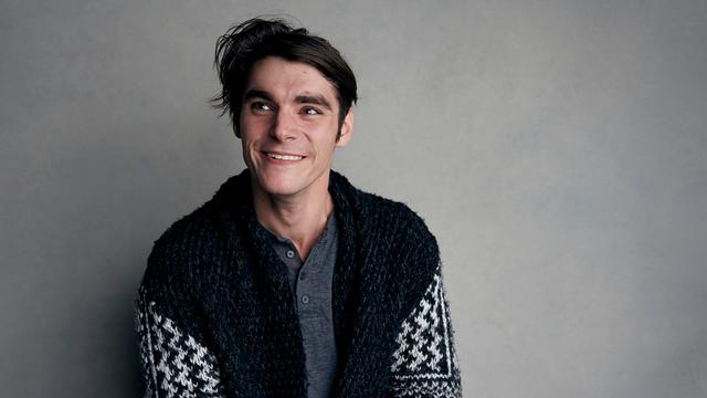 RJ Mitte sitzt auf einem Hocker vor einer grauen Wand. 