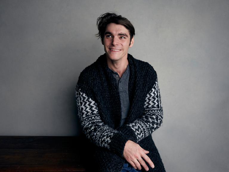 RJ Mitte sitzt auf einem Hocker vor einer grauen Wand. 