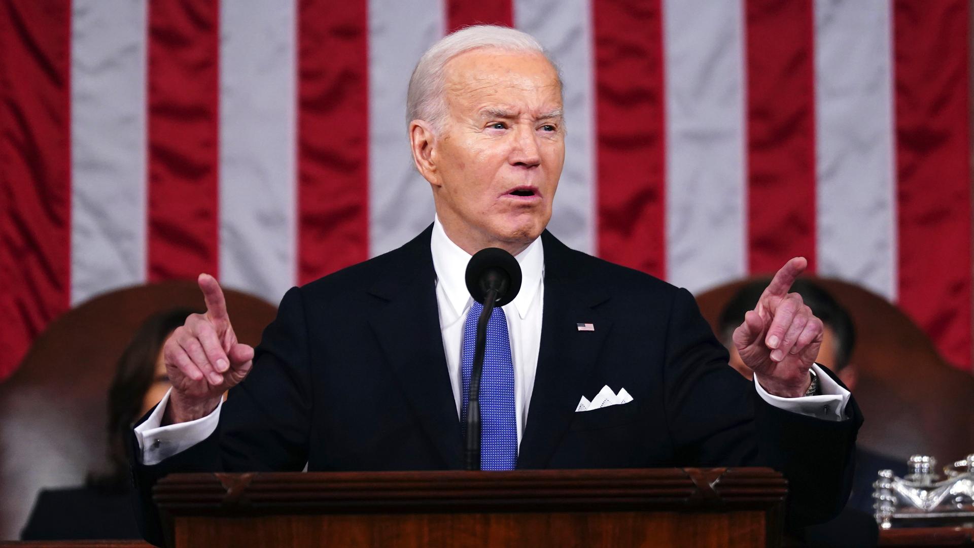 US-Präsident Joe Biden hält eine Rede. Er hebt beide Hände etwas hoch und streckt die beiden Zeige-Finger aus. Er steht vor einer US-amerikanischen Flagge.