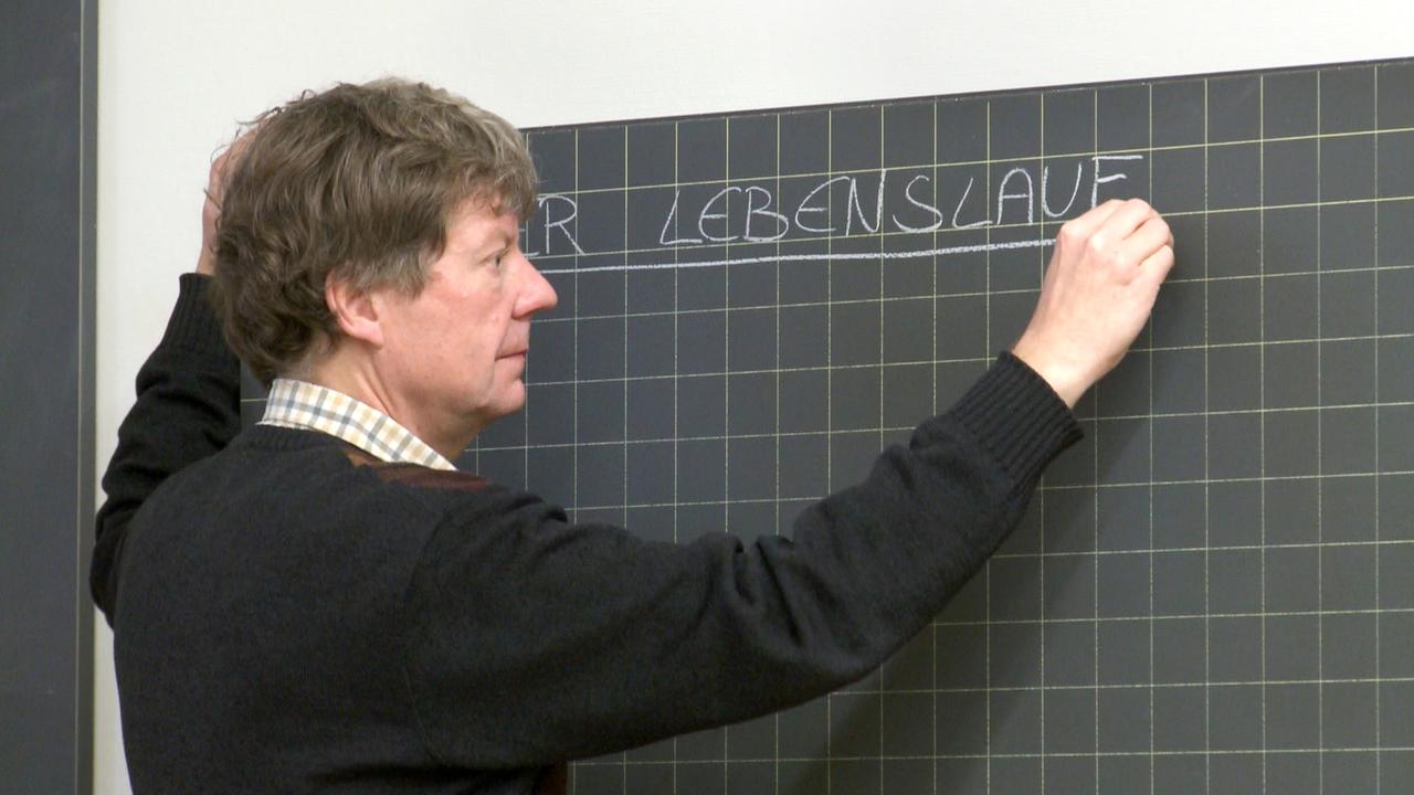 Christian Zingg steht vor einer Tafel und schreibt "Der Lebenslauf" mit Kreide.