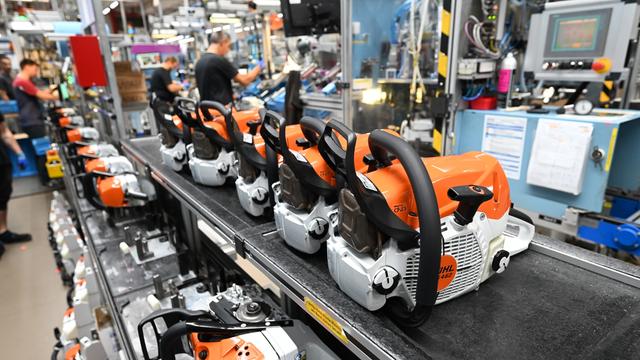 Im Stammwerk des Motorsägenherstellers Stihl werden an einem Band Sägen gefertigt. Die ikonischen orangenen Sägen liegen im Vordergrund auf einem Laufband. Im Hintergrund sind Arbeiter an Maschinen.