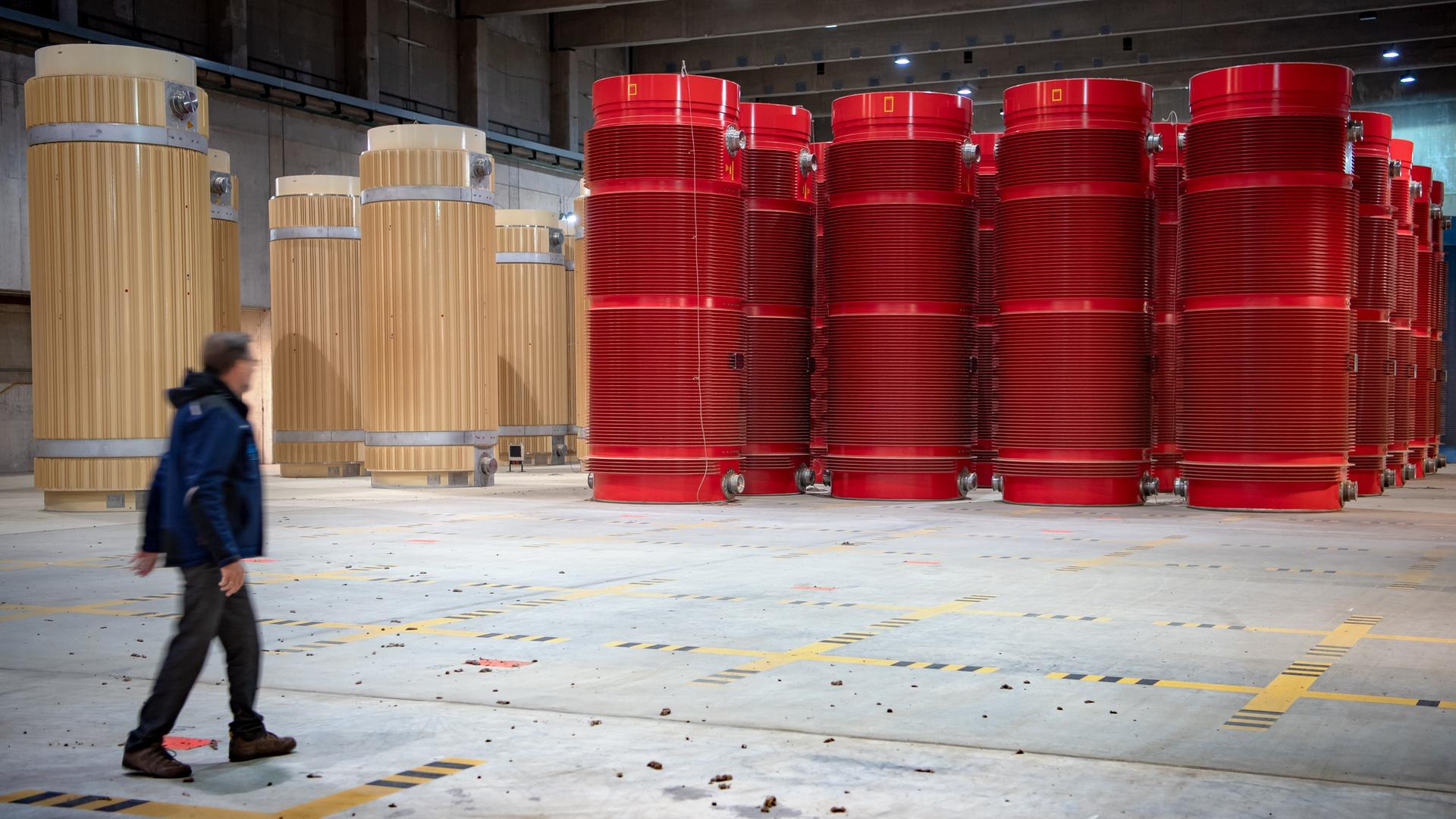 Eine Person läuft durch eine Halle, in der reihenweise tonnenförmige Behälter in beige und rot stehen, die Atommüll beinhalten. Eine Person läuft durch eine Halle, in der reihenweise tonnenförmige Behälter in beige und rot stehen, die Atommüll beinhalten.