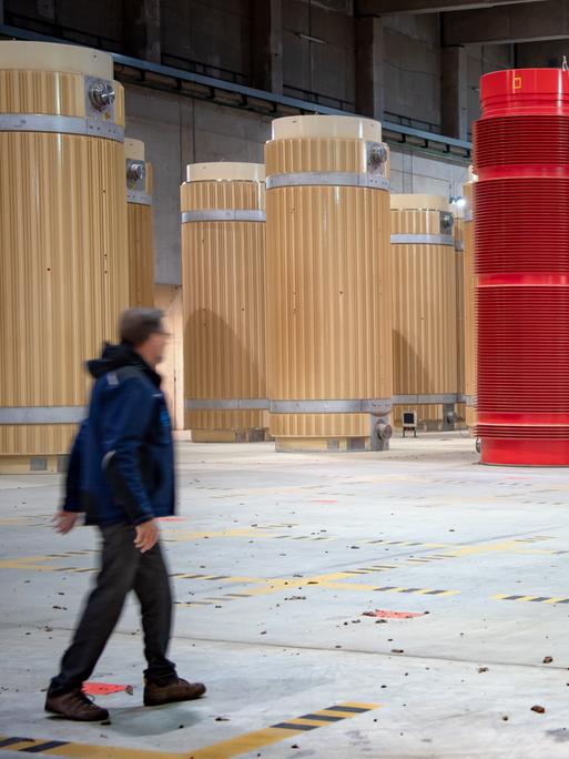 Eine Person läuft durch eine Halle, in der reihenweise tonnenförmige Behälter in beige und rot stehen, die Atommüll beinhalten.