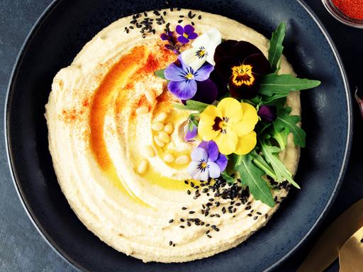 A beautifully plated bowl of creamy hummus garnished with olive oil, black sesame seeds, pine nuts, paprika, arugula, and vibrant edible flowers, served with pita bread on a dark textured background. Perfect as a banner image for food-related content.