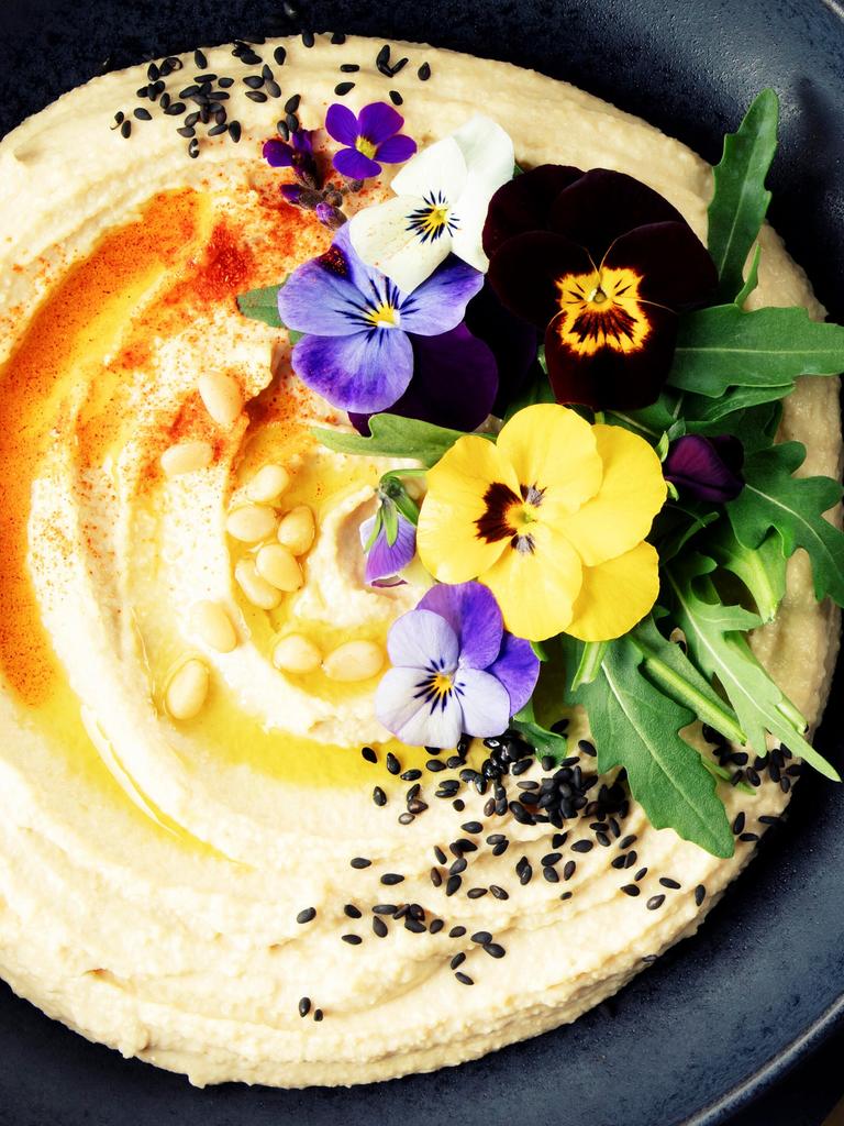 A beautifully plated bowl of creamy hummus garnished with olive oil, black sesame seeds, pine nuts, paprika, arugula, and vibrant edible flowers, served with pita bread on a dark textured background. Perfect as a banner image for food-related content.