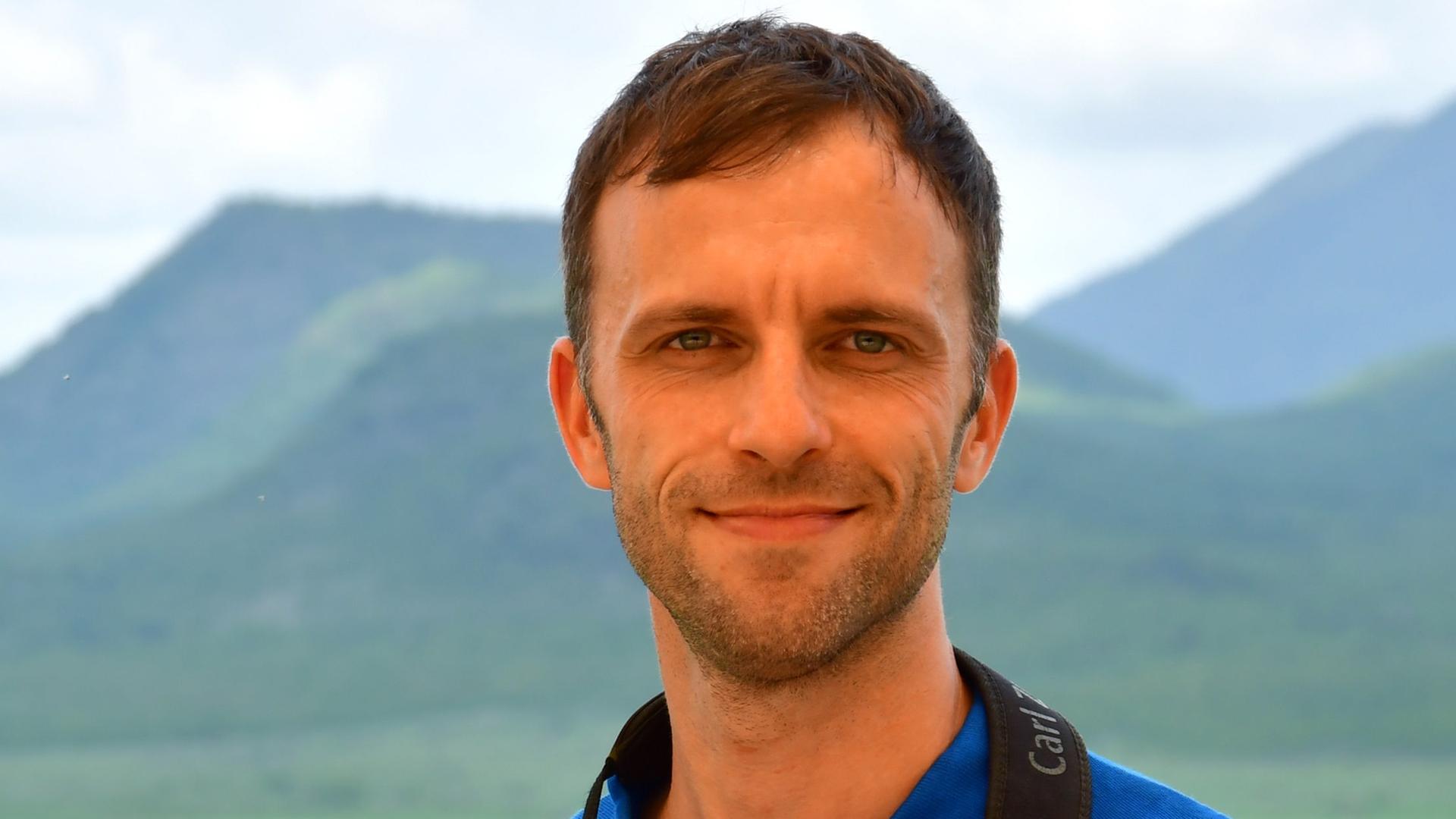 Moritz Klose steht vor einer im Hintergrund verschwommen sichtbaren Hügellandschaft. Er trägt ein blaues NABU-Poloshirt und hat ein Fernglas um den Hals gehängt, dass er mit seinen Händen festhält. Er guckt in die Kamera.