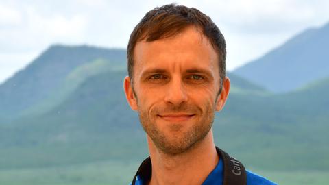 Moritz Klose steht vor einer im Hintergrund verschwommen sichtbaren Hügellandschaft. Er trägt ein blaues NABU-Poloshirt und hat ein Fernglas um den Hals gehängt, dass er mit seinen Händen festhält. Er guckt in die Kamera.