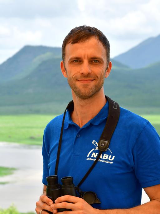 Moritz Klose steht vor einer im Hintergrund verschwommen sichtbaren Hügellandschaft. Er trägt ein blaues NABU-Poloshirt und hat ein Fernglas um den Hals gehängt, dass er mit seinen Händen festhält. Er guckt in die Kamera.
