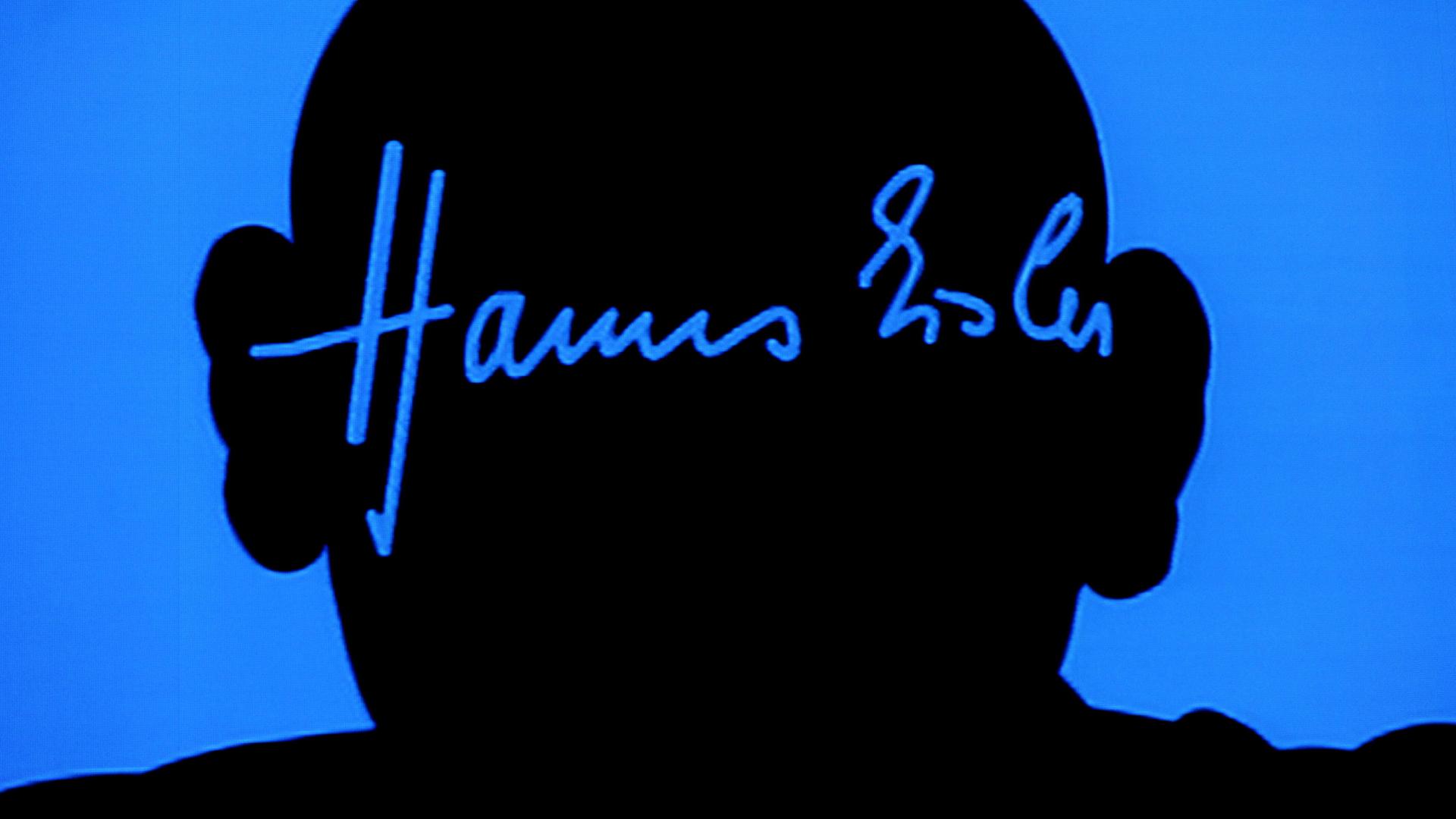 Der Schattenriss von Hanns Eisler ist an eine leuchtend blaue Wand geworfen, darin sein Autogramm in der blauen Farbe, die den Kopf umgibt. 