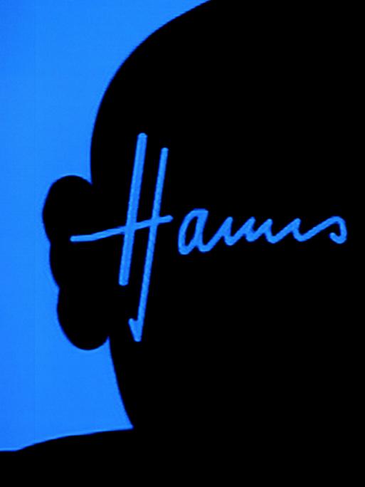 Der Schattenriss von Hanns Eisler ist an eine leuchtend blaue Wand geworfen, darin sein Autogramm in der blauen Farbe, die den Kopf umgibt. 