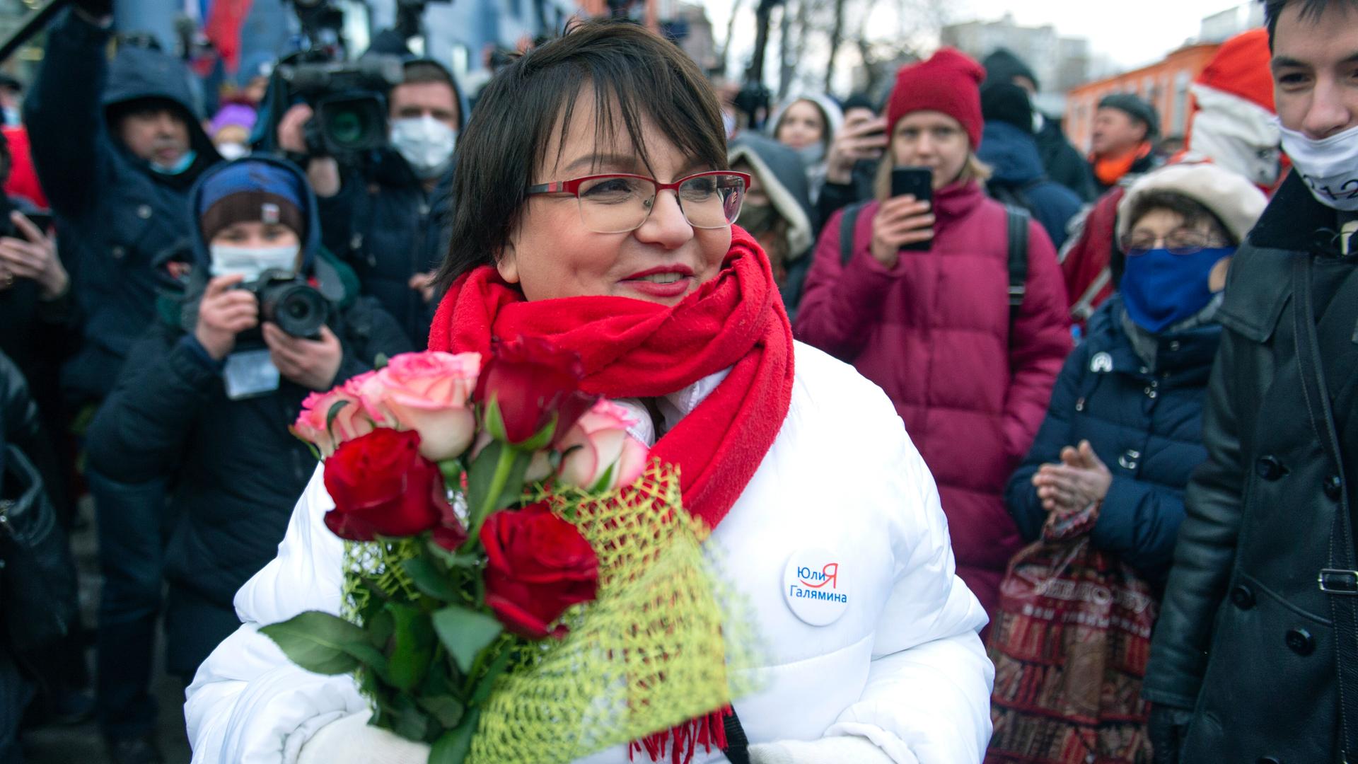 Yulia Galyamina steht lächelnd mit einem Blumenstrauß in Händen vor Pressevertretern auf der Straße.